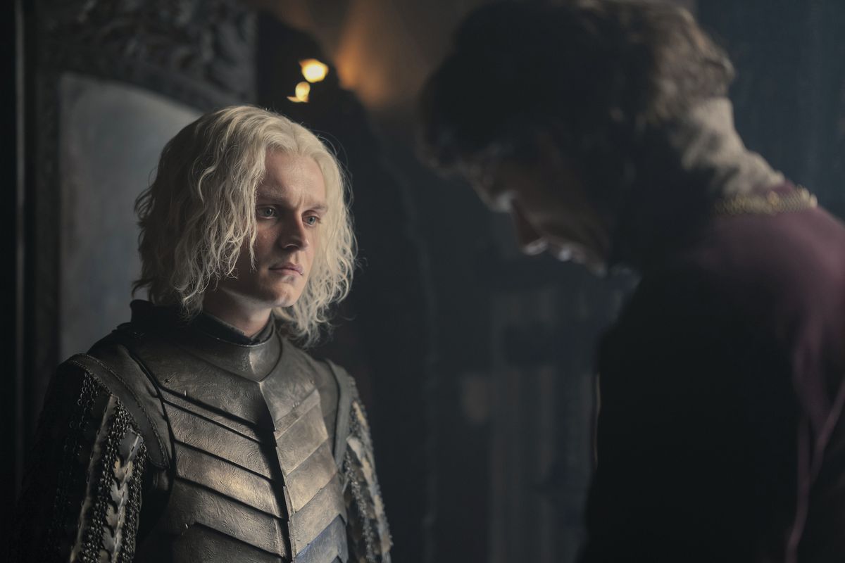 Tom-Glynn Carney como Aegon Targaryen em House of the Dragon, vestindo armadura e conversando com Larys Strong interpretado por Matthew Needham